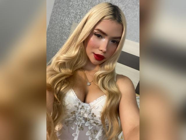 ChristieGarci has long blonde hair, red lipstick, and is wearing a white lace dress with floral patterns, showing her tits. She's looking at the camera with a slight smile.