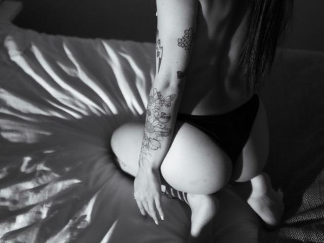 Fotografía en blanco y negro de MeliMuller, sin sostén, arrodillada en la cama, vistiendo ropa interior oscura, mostrando un brazo tatuado, glúteos y cabello largo y liso, de espaldas a la cámara, con una mano en la cama..