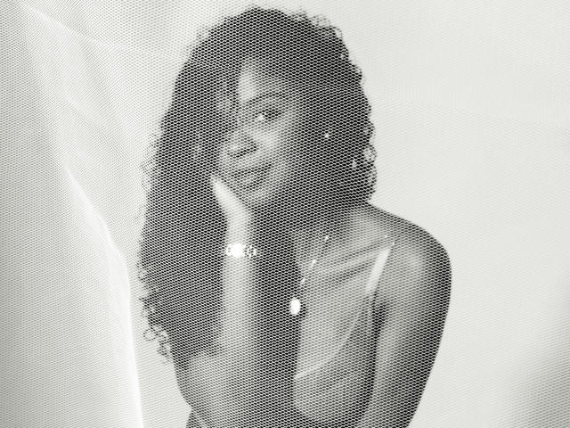 Black-and-white photo of LisaMendez with long curly hair, wearing a sheer top, covering her small tits with one hand, looking seductive, and wearing a necklace.