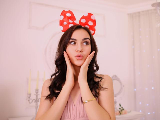 EricaWings com laço vermelho de bolinhas, lábios curvados, mãos nas bochechas, olhando de lado, vestindo um vestido rosa, cabelo castanho longo e ondulado, pulseira dourada, fundo branco suave, expressão fofa..