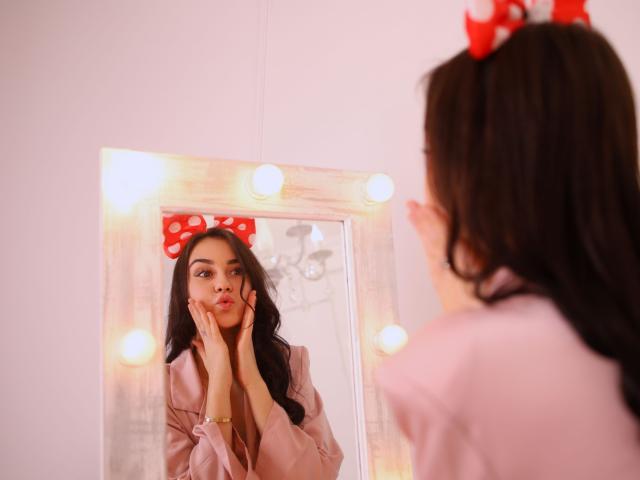 EricaWings met donker haar, een rode strik met stippen, een roze blouse, handen op de wangen, die in een spiegel kijkt met lichtgevende lampen, reflecterend, een zachtroze muur, een lichtjes fronsende uitdrukking, een mooi gezicht..