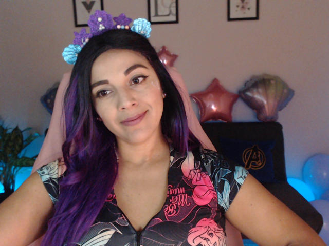 PrettyEllie with purple and black hair, wearing a blue and purple cat ear headband, smiling at the camera, in a floral shirt, with a slight pout on her ass.