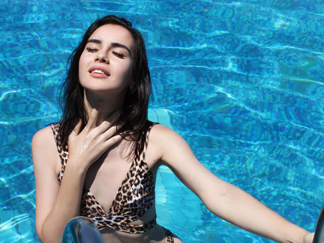 EricaWings mit nassen, schwarzen Haaren, einem Bikini-Oberteil mit Leopardenmuster, blauem Wasser, einer Hand am Hals, lächelnd, Wassertropfen auf der Haut, schwebend, halb geschlossene Augen, entspannt, sonniger Tag..