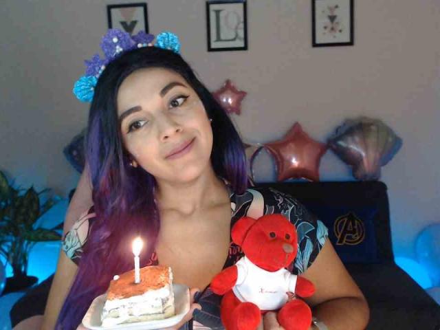 PrettyEllie with purple hair, wearing a blue headband, holding a cake with a candle, and a red teddy bear, smiling, in a cozy room with star balloons.