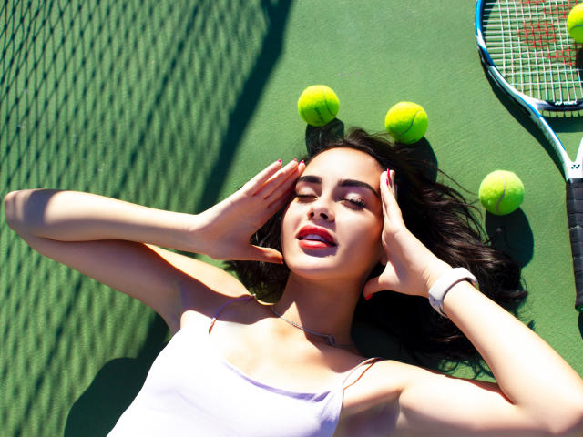 EricaWings ligt op een groene tennisbaan, in het zonlicht, met haar handen voor haar gezicht, rode lippen, een wit tanktopje, zwart haar, tennisballen en een racket, en draagt een witte polsband..