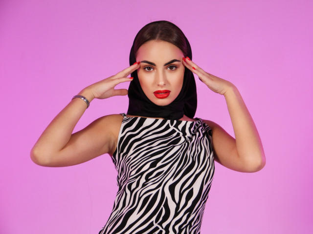 Melisa Wiesel tem lábios vermelhos, cabelo castanho escuro preso em um hijab preto, e está vestindo um vestido com estampa de zebra, com as mãos nos templos, contra um fundo rosa..