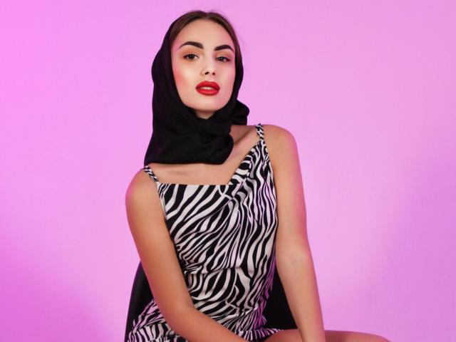 MelisaWiesel with red lips, wearing a black headscarf, zebra-pattern dress, sitting with legs apart, pink background, looking at camera with a slight smile, showing her tits.
