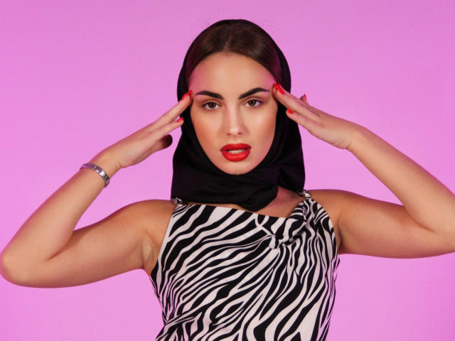 Melisa Wiesel, con las manos en las orejas, vestida con un hijab negro, un vestido de estampado de cebra, lápiz labial rojo, una pulsera, y un fondo rosa. Sus pechos son pequeños, y está mirando directamente a la cámara..