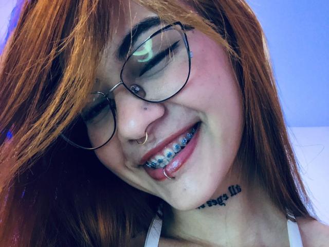 CieloSmith ha i capelli arancioni, occhiali, un anello al naso e una protesi dentale blu, sorride indossando una canottiera bianca, mostrando i denti inferiori e un anello al labbro, e ha un tatuaggio sotto il mento..
