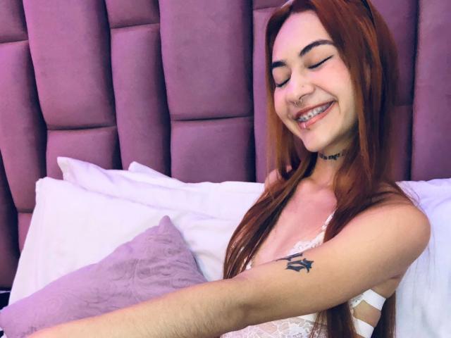 CieloSmith, con cabello rojo, ojos cerrados, sonriendo, vestida con lencería blanca de encaje, mostrando un tatuaje en su brazo, acostada en una cama con almohadas moradas..