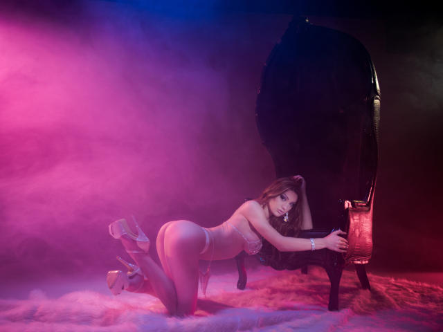 CataleyaLee está a cuatro patas, vestida con lencería y tacones altos, con las nalgas levantadas y los pechos pegados a una silla negra, rodeada de humo rosa y morado..