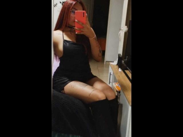 Alexandra Petit, com cabelo ruivo, vestindo um vestido preto, sentada em uma cama, tirando uma selfie no espelho com um celular vermelho, mostrando suas nádegas e tatuagens, em um quarto escuro..