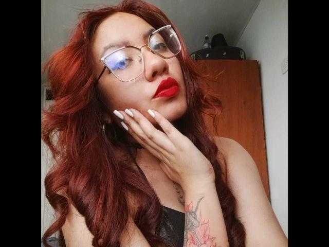 Alexandra Petit, com cabelo ruivo encaracolado, óculos, batom vermelho, unhas brancas, lingerie preta, tatuagem no pulso direito, mão no rosto, olhando para cima, sexy, confiante, atraente..