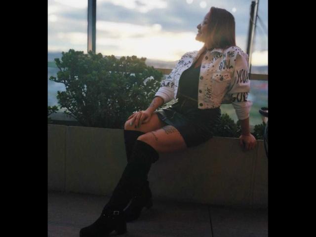 Alexandra Petit, portant une veste blanche à boutons et motifs, un short noir et des bottes montantes, assise sur une bordure en béton, montrant sa cuisse tatouée et regardant le coucher de soleil..