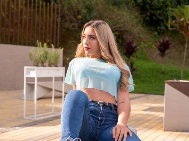EmmaMillers with blonde hair, blue crop top, and blue jeans, sitting on a wooden deck, showing her tits and a tattoo on her left arm. EmmaMillers looks to the side.