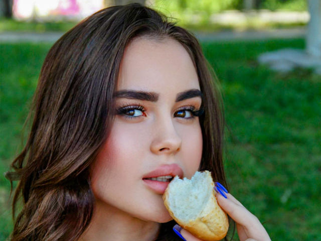 EricaWings, met bruin golvend haar, blauwe nagels, die een halve bagel eet, heeft roze wangen, bruine ogen en volle, roze lippen, in een groene, open omgeving..