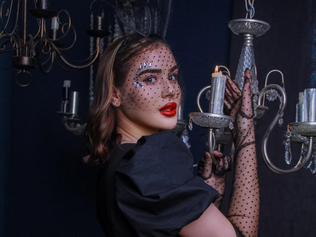 EricaWings con labios rojos, una máscara de lunares negros y un vestido negro, sosteniendo una vela encendida, usando guantes de lunares negros, de pie cerca de elaborados candelabros de plata..