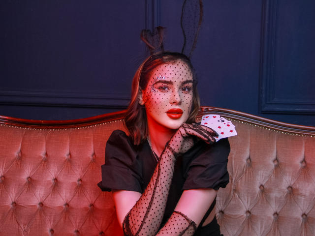 EricaWings with black polka-dot gloves, holding a heart-faced card, wearing a black shirt, and black bunny ears, sitting on a tufted beige couch, red light on face.