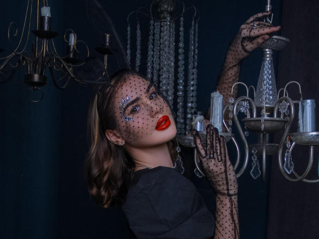 EricaWings, avec des lèvres rouges, des gants en maille noire et un fard à paupières scintillant, tenant un lustre en argent, portant un haut noir, regardant vers le haut avec une expression séduisante..
