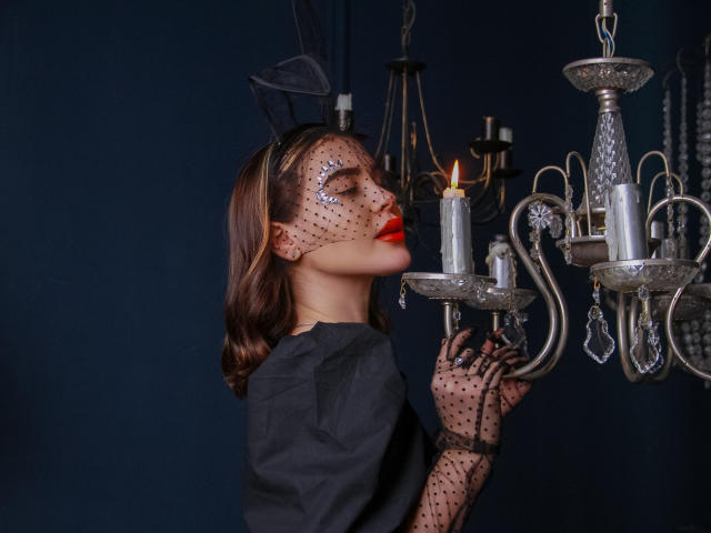 EricaWings, con labios rojos, vestido negro y máscara de lunares, olfatea una vela encendida en un candelabro de plata, vistiendo guantes de encaje negro, sobre un fondo azul oscuro..