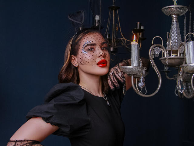 EricaWings avec rouge à lèvres rouge, robe noire, manches bouffantes, masque transparent à pois et pièce de tête noire, tenant un chandelier en argent, regardant vers le haut, dans une photo de style victorien, sombre et atmosphérique..