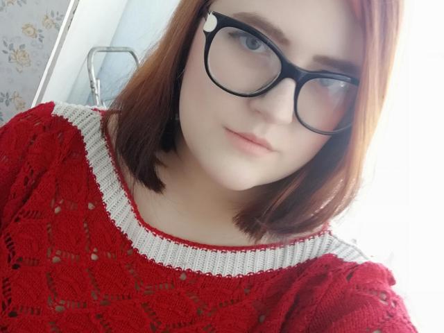 Melissa, con i capelli rossi, gli occhiali neri e la pelle chiara, indossa un maglione lavorato a maglia rosso e bianco, e guarda la fotocamera con un leggero sorriso. Il seno non è visibile..