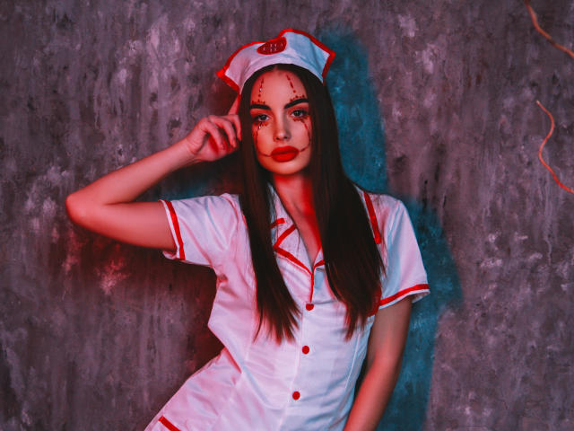 MelisaWiesel in einer Krankenschwesteruniform in Weiß und Rot, mit einer Krankenschwestermütze mit roten Kreuzen, dunklem, glattem Haar, roter Gesichtsfarbe und rotem Lippenstift, posiert vor einer rauen Wand..