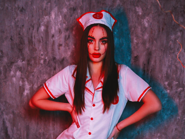 Melisa Wiesel, com longos cabelos castanhos, vestindo um uniforme de enfermeira vermelho e branco, com as mãos nos quadris, batom vermelho, sangue no rosto, chapéu de enfermeira branco, cruz vermelha, botões vermelhos, detalhes vermelhos..