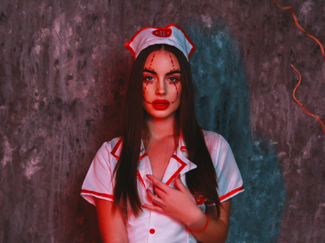 MelisaWiesel with bloody face, wearing a white nurse outfit with red trim, red lipstick, and a nurse cap, standing against a textured wall, hand on chest.