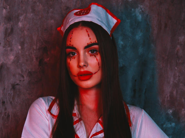 MelisaWiesel in a white nurse hat with red trim, bloody face makeup, and red lipstick, wearing a white nurse uniform with red accents, looking serious.