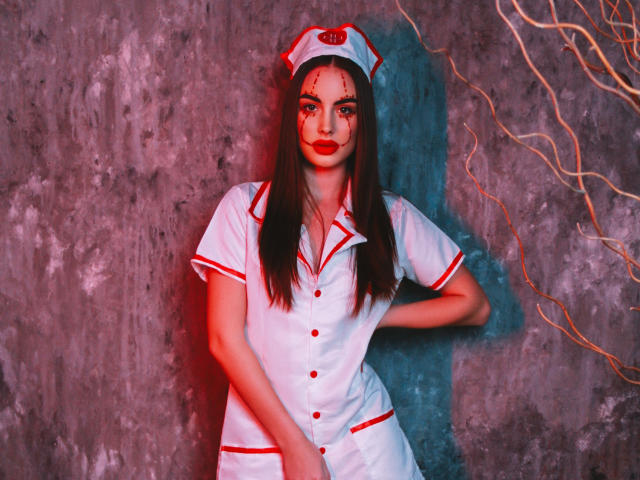Melisa Wiesel vestida com um uniforme de enfermeira branco com detalhes vermelhos e um chapéu, com sangue no rosto, em pé contra uma parede cinza com galhos retorcidos, com as mãos na cintura..