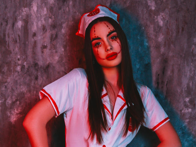 Melisa Wiesel, con cabello largo y oscuro, vestida con un uniforme blanco de enfermera, lápiz labial rojo y sangre en la cara, de pie frente a una pared texturizada con iluminación roja y azul..
