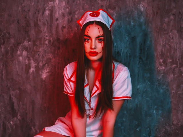 Melisa Wiesel met een bloedig gezicht, rode lippenstift en lang donker haar, gekleed in een witte verpleegsteroutfit met rode accenten, zittend tegen een donkere, gestructureerde muur, met rood en blauw licht..