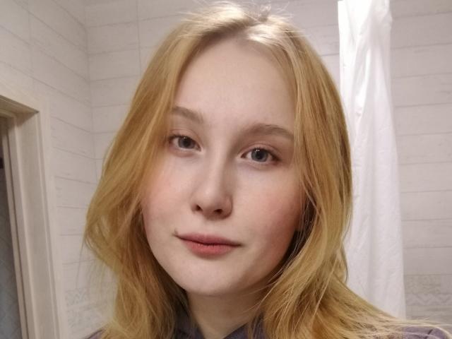 AnikaLipps a les cheveux blond clair, une peau claire et des yeux bleus. Elle regarde l'appareil photo avec un léger sourire, et porte une chemise grise, dans une salle de bain..