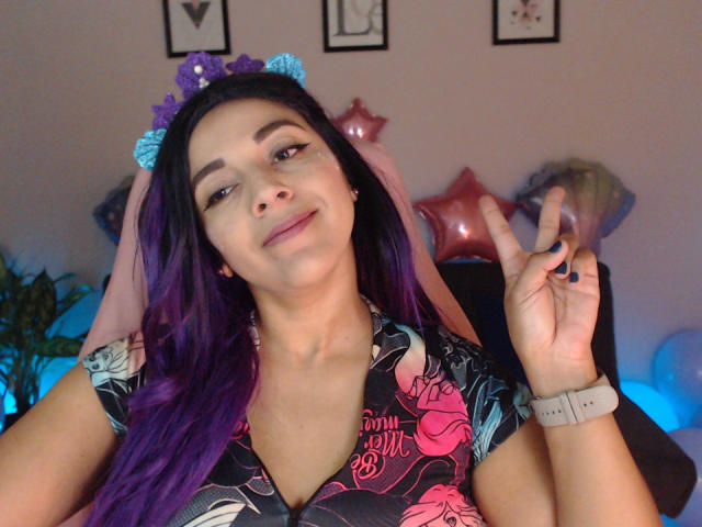 PrettyEllie with purple hair, blue butterfly headband, pink veil, floral shirt, and pink wristband, making a peace sign, smiling, showing off dark nail polish.