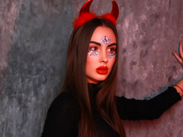 Melisa Wiesel, com chifres vermelhos de diabo, vestindo um suéter preto, batom vermelho e maquiagem brilhante nos olhos, tocando uma parede cinza com textura. Seu cabelo castanho é longo e liso..