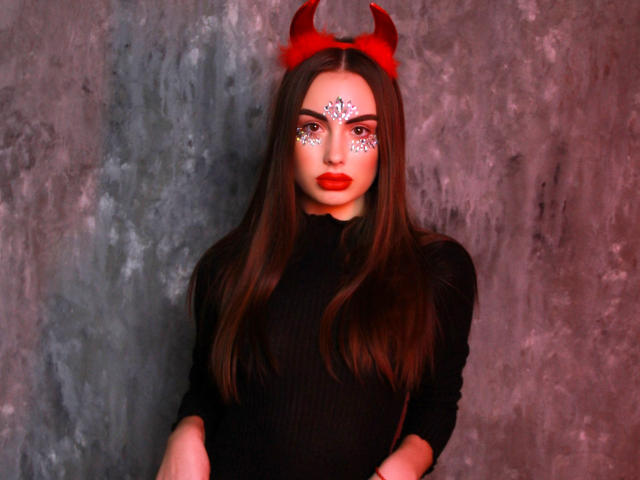 MelisaWiesel with red devil horns, black ribbed turtleneck, red lipstick, white diamond eye makeup, long brown hair, grey textured wall, red lighting, sexy, attractive, confident.