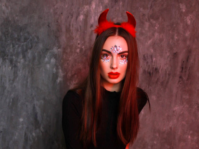 Melisa Wiesel met rode hoorns, lang bruin haar, een zwarte top, rode gezichtsbeschildering, glitter en rode lipstick, met een serieuze blik. Haar borsten zijn bedekt door de zwarte top..