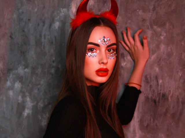 MelisaWiesel with red devil horns, sparkly face paint, and red lipstick, wearing a black top, touching a gray wall with her right hand. Long brown hair, red nail polish.