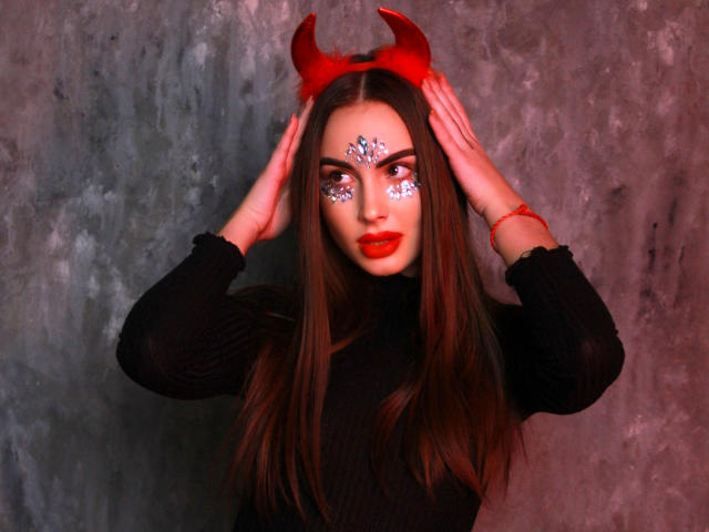 Melisa Wiesel con corna da diavolo rosse, maglione nero a collo alto, rossetto rosso, trucco bianco sul viso, mani sulla testa, lunghi capelli castani, parete grigia con texture, braccialetto rosso, che guarda la fotocamera..