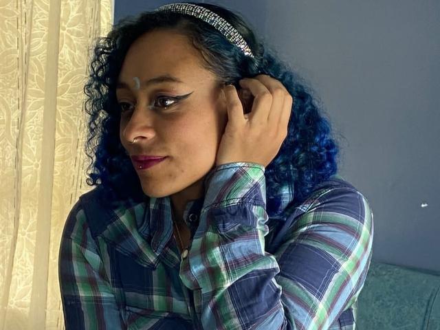 CataXBlue with blue curly hair, wearing a plaid shirt, silver headband, and purple lipstick, has a white mark on her forehead, touching her ear with her hand.