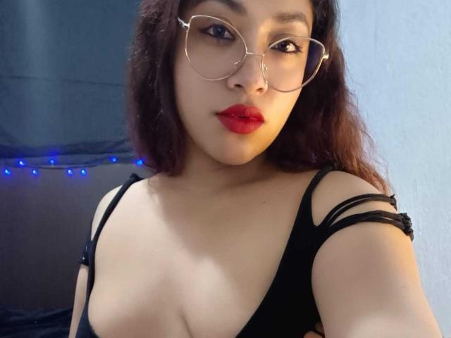 AmelyKent has wavy brown hair, red lipstick, and wears glasses with a black top showing her tits and strappy sleeves. She's looking at the camera with a slight smile.