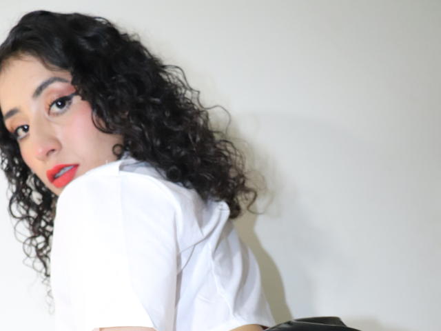Valerie Masse, con i capelli neri ricci, il rossetto rosso e la camicia bianca, guarda oltre la spalla, mostrando il seno e un accenno del sedere..