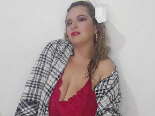 NaugthyCurvy avec rouge à lèvres rouge, fard à paupières rose, nœud blanc, cheveux bruns ondulés, boucles d'oreilles dorées et haut à carreaux noir et blanc, souriant à la caméra..