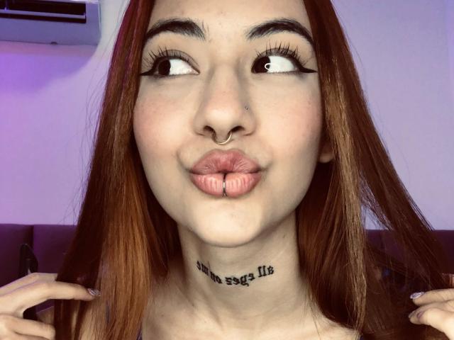 AlixLow mit schwarzen Haaren, Brille und gebräunter Haut, oben ohne, zeigt ihr Gesäß in engen weißen Hosen, lehnt sich in einer roten Küche an, Tattoo auf dem Rücken..