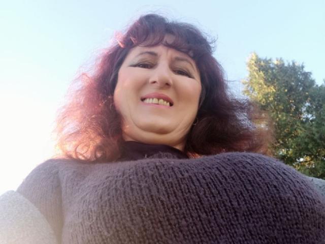 BellisimaAh mit lockigem, rotbraunem Haar, lächelnd in die Kamera, trägt einen strukturierten, grauen Pullover, hat große Brüste und einen schwarzen Rollkragenpullover. Das Foto wurde aus einem niedrigen Winkel im Sonnenlicht aufgenommen..
