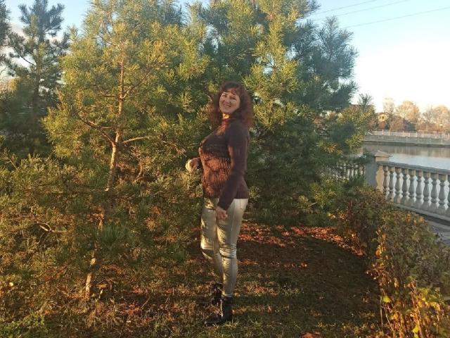 BellisimaAh stands outdoors, smiling, with her small tits visible under a sheer purple top, wearing tight, light-green pants and black boots, in front of a green pine tree.