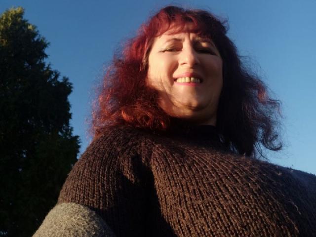 BellisimaAh mit roten, lockigen Haaren, lächelnd in die Kamera, trägt einen braunen Strickpullover, wobei ihre Brüste leicht sichtbar sind, vor einem klaren, blauen Himmel..