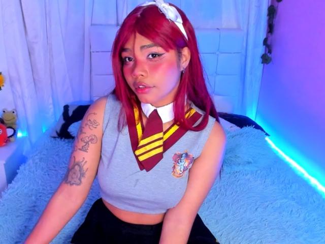 AnniieeBell, com cabelo ruivo, vestindo uma blusa cinza sem mangas com gravata listrada em amarelo e vermelho, calças pretas, uma faixa de cabelo branca, tatuagens no braço esquerdo, sentada em uma cama azul e fofa..
