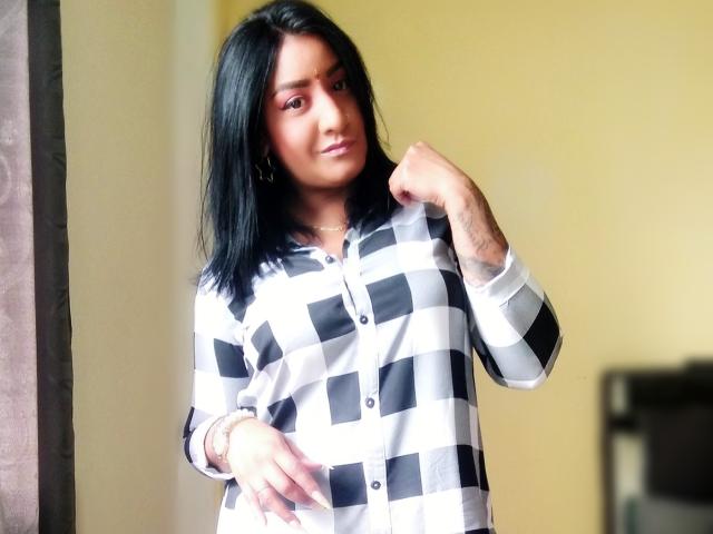 Nahirr with black hair, wearing a black-and-white checkered shirt, standing, right hand near face, left hand on stomach, subtle tattoo on right wrist, soft yellow background.
